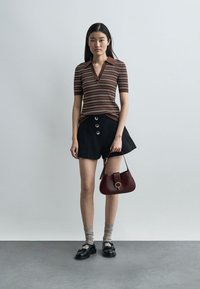 Frau steht und trägt ein braun gestreiftes Poloshirt, schwarze Shorts mit Knöpfen, graue Socken, schwarze Schuhe und hält eine kleine dunkelrote Handtasche.