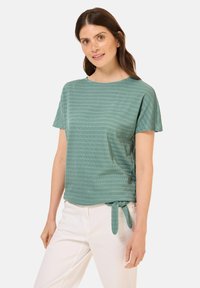 Tealová krátká halenka s kulatým výstřihem, texturovanými pruhy a uzlem na boku. V kombinaci s bílými kalhotami, které mají uvolněný střih.