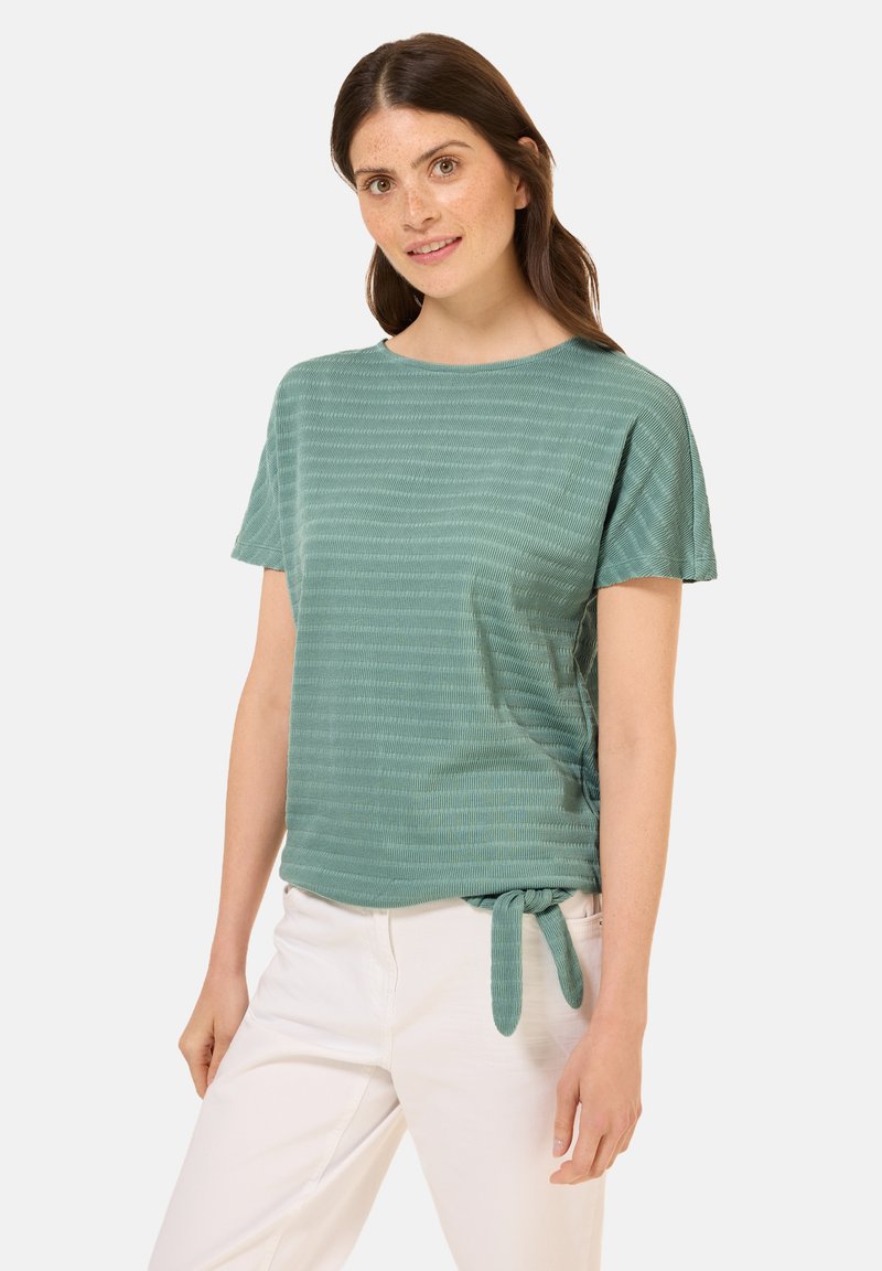 Tealová krátká halenka s kulatým výstřihem, texturovanými pruhy a uzlem na boku. V kombinaci s bílými kalhotami, které mají uvolněný střih.