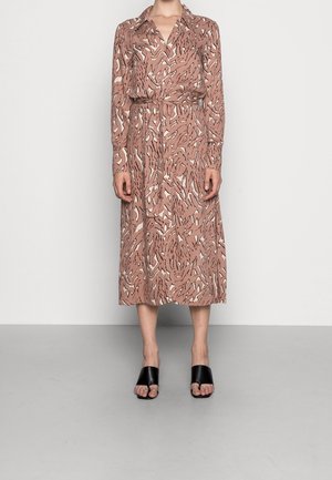 Frau trägt ein langärmeliges, knopfgeschlossenes Midikleid mit braunem, abstraktem Muster und schwarze, offene, mit Absatz versehene Maultiere, steht vor einer schlichten Wand.