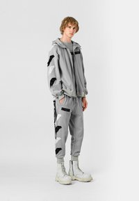 Chándal gris con diseños de alas en negro y blanco. Incluye una sudadera con cremallera y pantalones ajustados. Estilizado con botas de combate blancas.