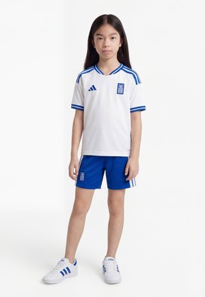 Enfant portant un uniforme de football blanc et bleu avec un emblème grec, debout les bras détendus contre un fond blanc.