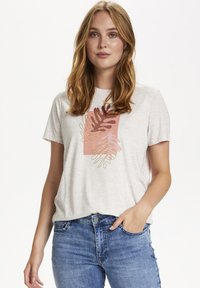 Jauna sieviete ar sarkaniem matiem valkā bēšas krāsas īsām piedurknēm krekls ar abstraktu lapu rakstu un zila džinsu, roka kabatā, uz balta fona.