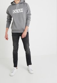 Felpa grigia con un logo bianco sul petto, abbinata a jeans neri e sneakers bianche. Presenta un cappuccio con cordino e polsini a coste.
