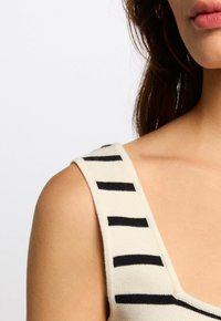 Débardeur crème avec des rayures noires, présentant une texture côtelée et des bretelles larges. L'encolure a une forme légèrement arrondie, mettant en valeur le design.