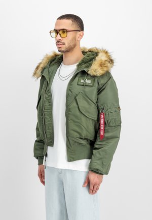 Mand iført grøn pelsforet hættejakke med rød mærkat, hvid skjorte, lyse jeans og gultonede briller, med blik mod venstre.