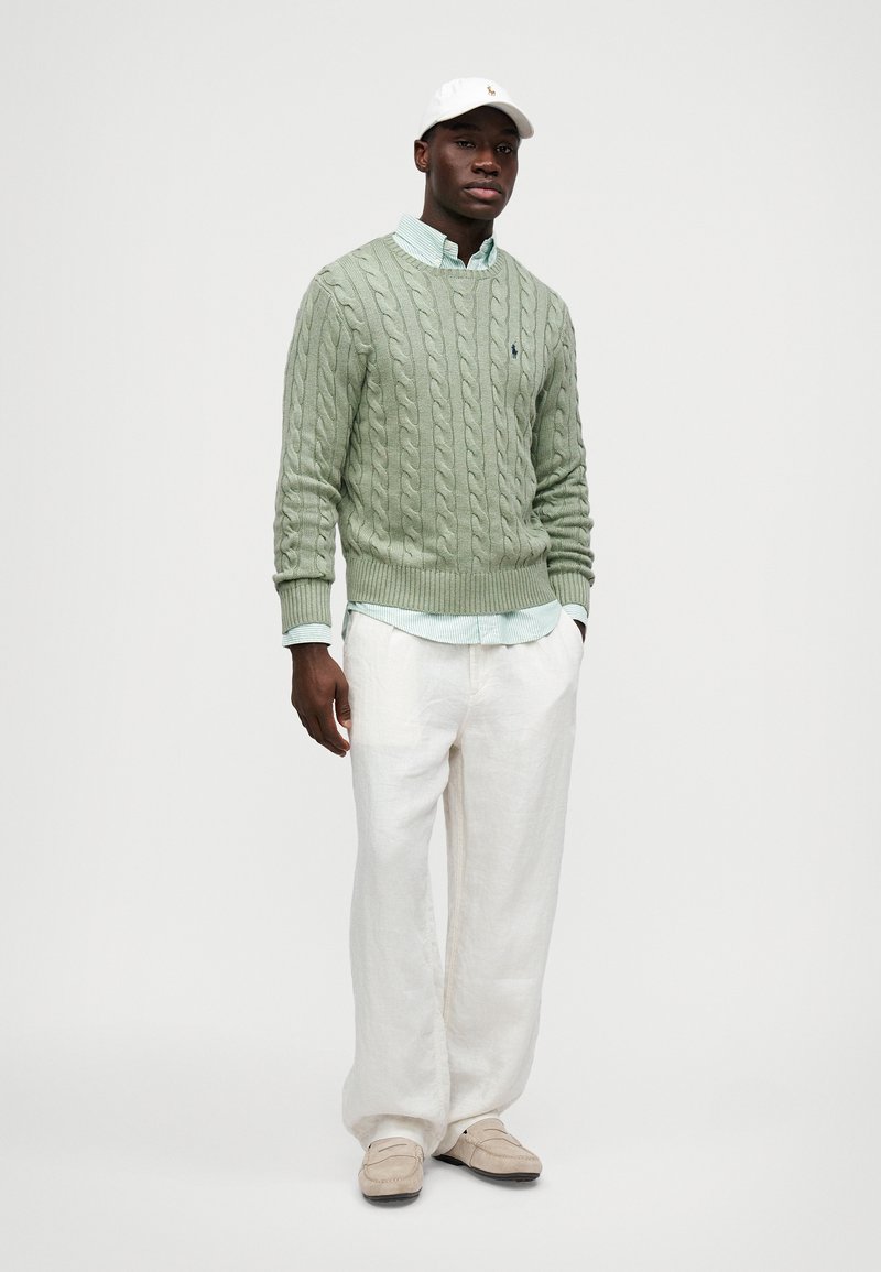 Suéter verde de punto trenzado sobre una camisa de cuello azul claro, combinado con pantalones de lino blancos y zapatos slip-on de color marrón claro. Gorra blanca.