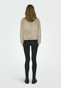 Beige, fuskpälsjacka med en kort design, visad bakifrån, tillsammans med mörka jeans och svarta ankelboots.
