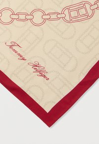 Hedvábný šátek s krémovým pozadím a červenými řetězovými vzory. Obšitý červeným lemem s nápisem "Tommy Hilfiger" psaným kurzívou v dolním rohu.