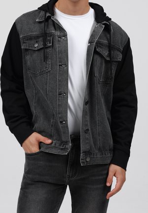 Mand iført en sort og grå denimjakke med sorte ærmer og hætte over en hvid t-shirt, med den ene hånd i lommen.
