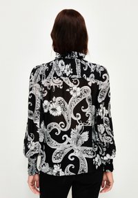 Schwarze Bluse mit floralem und paisleymuster in Weiß, langen Ärmeln und einem rüschierten Ausschnitt, aus einem leichten, semi-transparenten Stoff.