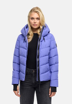 Blonde Frau, die eine helllila gepolsterte Kapuzenjacke über einem schwarzen Top und dunklen Jeans trägt, vor einem einfarbigen Hintergrund stehend.