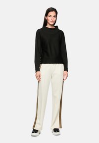 Schwarzes, kurzes Sweatshirt mit hohem Kragen, kombiniert mit weißen Hosen, die braune und goldene Rennstreifen an den Seiten haben. Schwarze Sneaker.