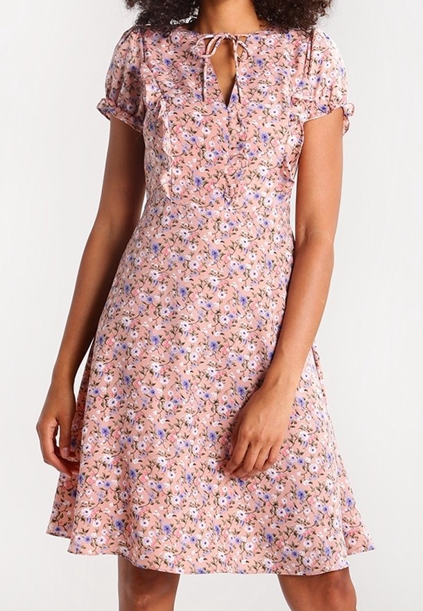 Femme portant une robe rose à manches courtes à motifs floraux avec un décolleté en forme de goutte noué avec un petit nœud.