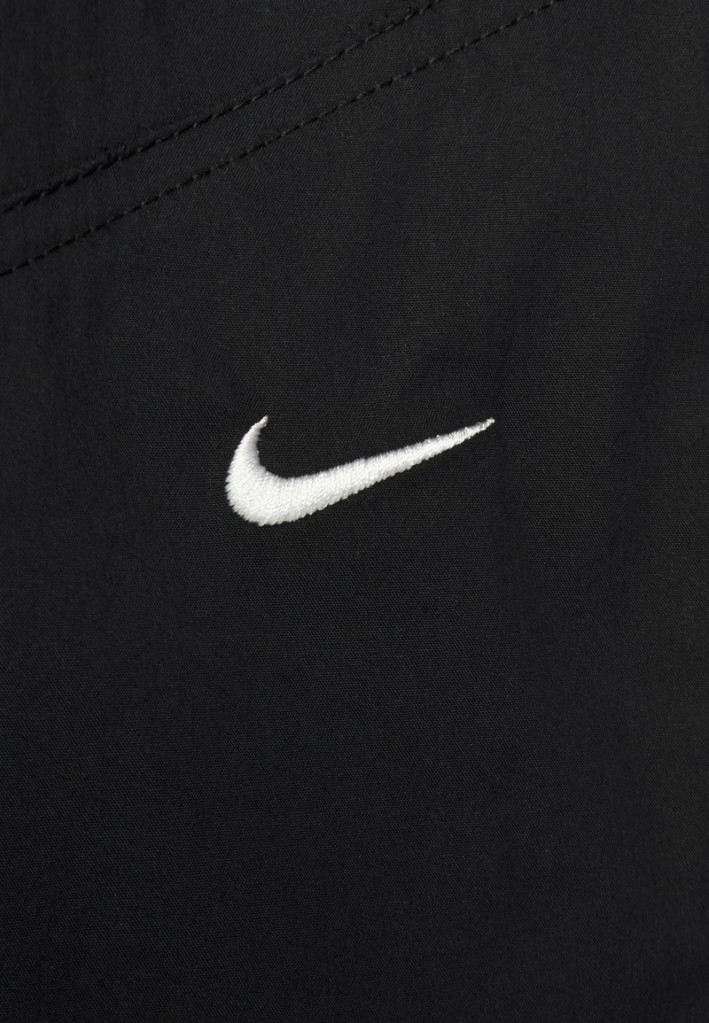 Weißes Nike-Swoosh-Logo auf schwarzem Stoff gestickt, mit sichtbarem diagonalen Stickdetail.