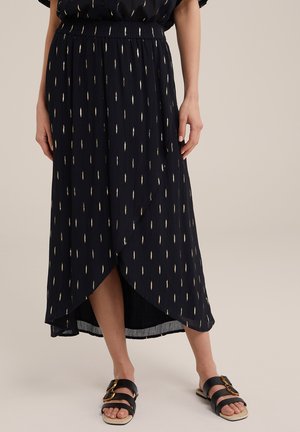 Mørk marineblå omslagsnederdel med metallic gyldent bladmønster, elastisk taljebånd og sideslids. Kombineret med sorte slidesandaler med gyldne detaljer.