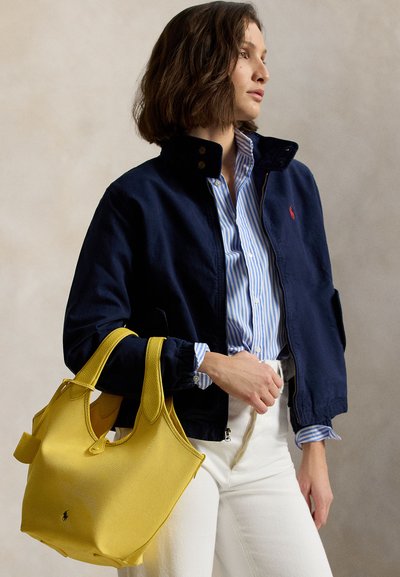 Γυναίκα με ναυτικό σακάκι και μπλε ριγέ πουκάμισο, κρατώντας μία κίτρινη τσάντα ώμου, φορώντας λευκό παντελόνι μπροστά σε ουδέτερο φόντο.