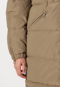 Veste rembourrée de couleur brun discret avec poche zippée, poignets élastiques et texture légère, idéale pour une tenue décontractée par temps frais.