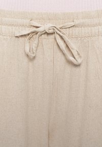 Smėlinės spalvos medvilninės kelnės su elastinga juosmens juostele ir raišteliu priekyje. Medžiaga minkšta, šiek tiek tekstūruota, matomos siūlės.