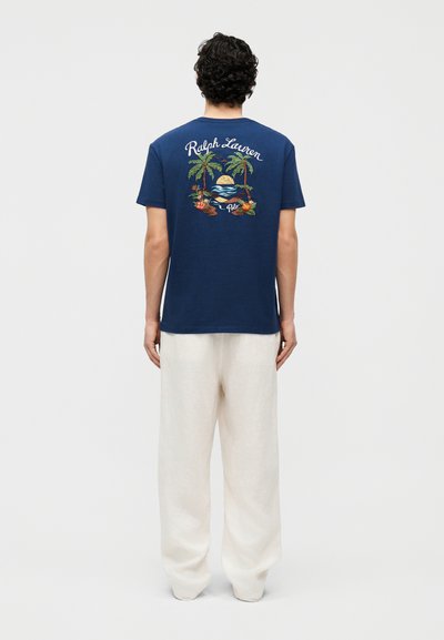 Άνδρας που στέκεται με την πλάτη γυρισμένη, φορά ένα μπλε ναυτικό μπλουζάκι με εικόνα τροπικού ηλιοβασιλέματος και το κείμενο "Ralph Lauren Polo" στο πίσω μέρος, καθώς και άνετο λευκό παντελόνι.