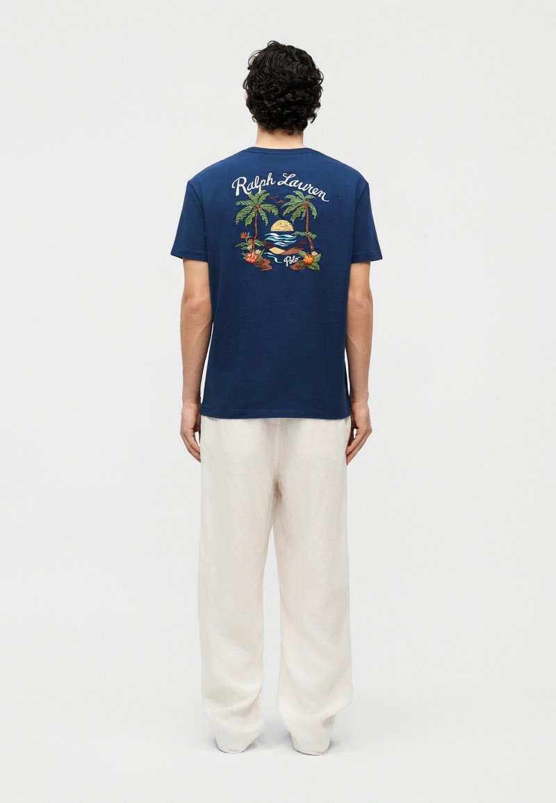 Man die met de rug naar de camera staat, draagt een marineblauw T-shirt met een tropische zonsondergang en de tekst "Ralph Lauren Polo" op de achterkant, en losse witte broek.