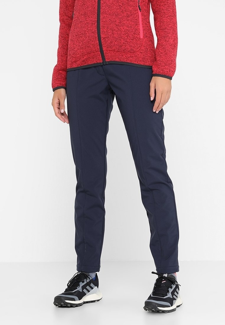 Personne portant un pantalon slim bleu marine, des baskets noires et blanches, et une veste zippée rouge, les mains détendues le long du corps.