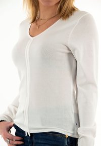 Valkoinen pitkähihainen neulepaita V-pääkkeen ja etuvetoketjuyksityiskohdalla, jossa on teksturoitu pinta ja slim fit -malli, yhdistettynä sinisiin farkkuihin.