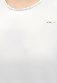 Κοντινό πλάνο ενός ατόμου που φοράει απλή λευκή μπλούζα με στρογγυλή λαιμόκοψη και μικρό λευκό κεντητό λογότυπο κροκόδειλου στο αριστερό στήθος.