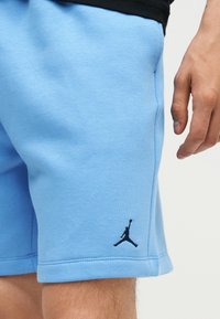 Ανοιχτό μπλε σορτς με μαύρο λογότυπο Jumpman στο αριστερό πόδι, που φοριέται από άτομο με ορατό χέρι και μέρος μαύρου πουκαμίσου.