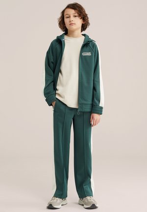 Jeune personne portant un survêtement vert foncé avec des bandes blanches, un t-shirt beige et des baskets grises, debout devant un fond uni.