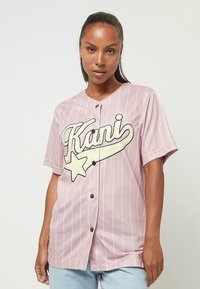 Rosa Baseballtrikot mit vertikalen weißen Streifen, das ein großes schwarzes und cremefarbenes Logo zeigt. Kurze Ärmel und Knopfverschluss. Weicher Stoff.