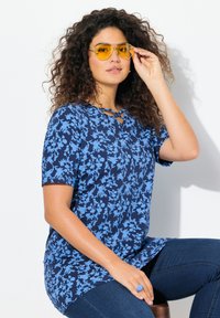 Camiseta de manga corta con patrón floral azul, que presenta un escote en V y detalles de tiras. Combinada con gafas de sol amarillas y un anillo azul.