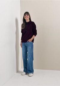 Felpa bordeaux scuro con tasca frontale e cordini, abbinata a jeans a gamba larga blu chiaro e sneaker bianche. Look semplice e casual.