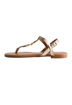 Płaska beżowa sandałka z metalicznym złotym paskiem typu T, regulowaną klamrą oraz dekoracyjnym łańcuszkiem na białym tle.