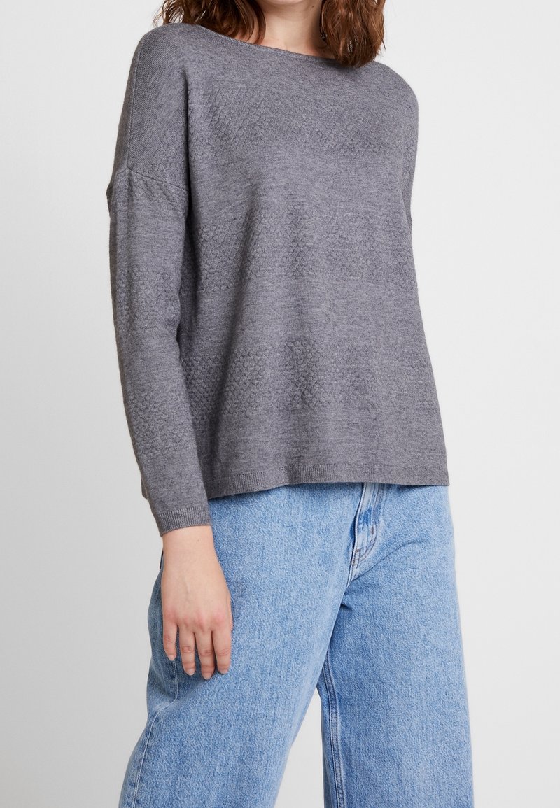 Femme portant un pull gris texturé à manches longues et un jean en denim bleu clair taille haute, debout devant un fond uni.