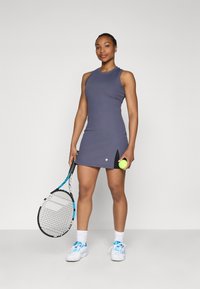 Tumehall hallituseta tennisekleit, millel on külgpertsed, hoides kollast tennisepalli ja reketit. Valged spordijalatsid ja sokid.