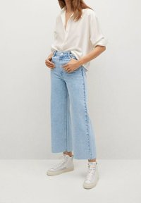 Personne portant un chemisier blanc boutonné, un jean taille haute à jambes larges bleu clair et des baskets montantes blanches, sur fond uni.