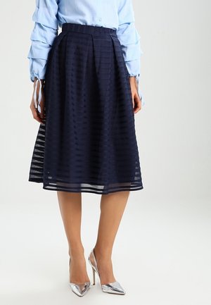 Femme portant une jupe plissée bleu marine, une blouse bleu clair à manches longues avec des poignets noués et des chaussures à talons hauts argentées.