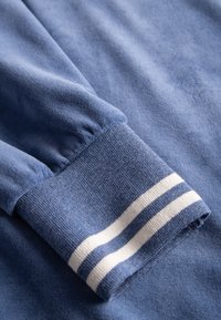 Blauer Pullover mit einer strukturierten Stoffoberfläche und geripptem Bündchen, akzentuiert durch weiße Streifen. Das Bündchen ist gerafft, was das Design unterstreicht.