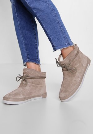 Personne portant un jean bleu et des bottines beiges en daim avec des lacets noués en nœud, debout sur un fond clair uni.