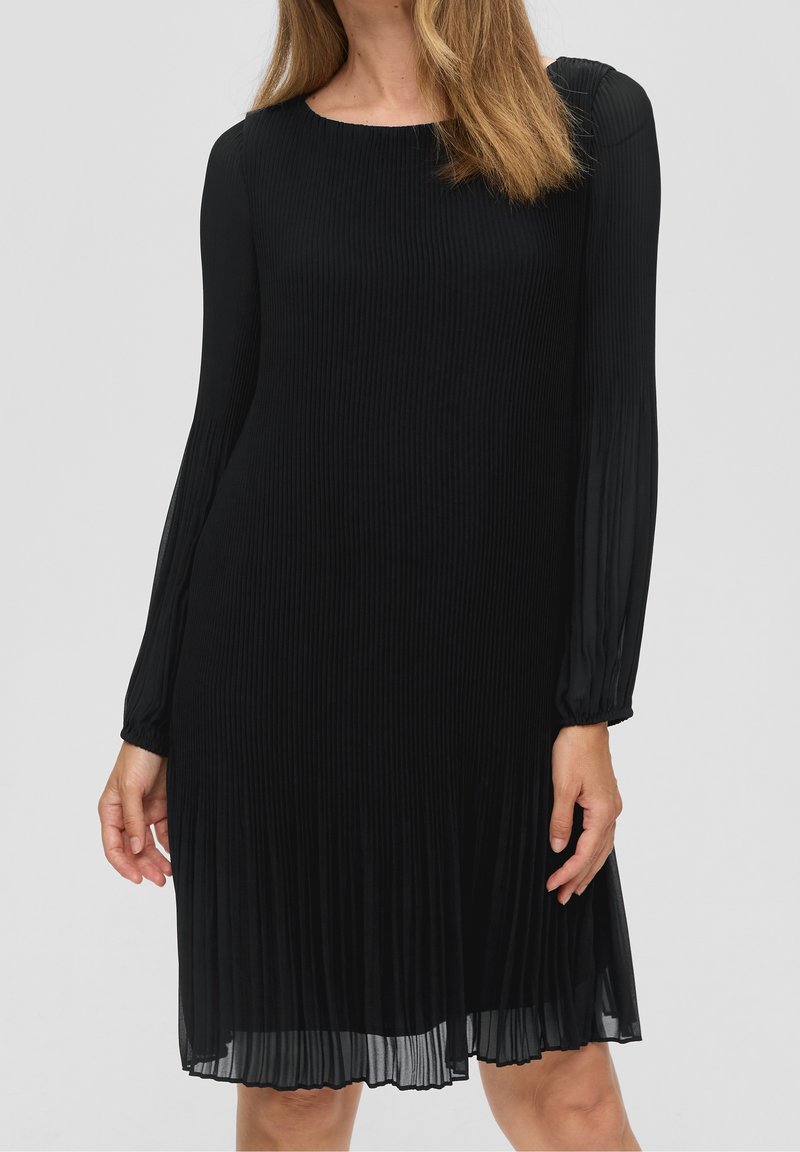 Femme portant une robe noire plissée à manches longues, longueur genoux, avec un col rond sur fond uni.