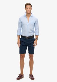 Camisa de lino azul claro de botonadura con mangas remangadas, pantalones cortos marinos y zapatos de barco de cuero marrón. El modelo está de pie en posición vertical frente a un fondo liso.