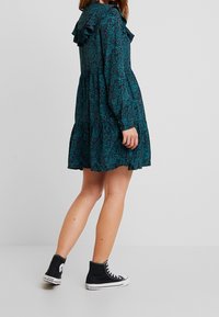 Žena v tyrkysovém vzorovaném dlouhém šatě s volány a černých vysokých teniskách Converse, stojící na pozadí jednotného světlého pozadí.