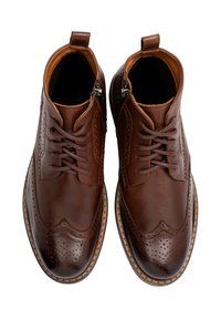Brązowe skórzane botki za kostkę z fasonem wingtip, z zapięciem na sznurowadła i zamkiem błyskawicznym. Posiadają dekoracyjne perforacje oraz kontrastującą jasną podeszwę.