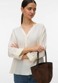 Femme aux cheveux foncés attachés en queue de cheval, portant un chemisier blanc ample et un jean, tenant un sac à main en suède marron foncé par ses anses.