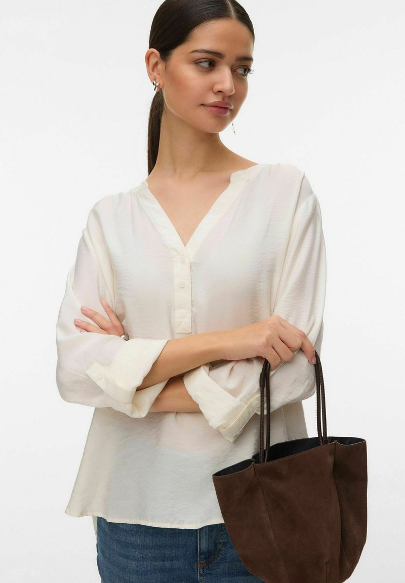 Femme aux cheveux foncés attachés en queue de cheval, portant un chemisier blanc ample et un jean, tenant un sac à main en suède marron foncé par ses anses.