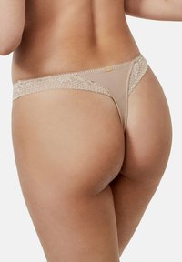 String en dentelle dans une teinte beige neutre, présentant un design en maille transparente et des accents à motifs complexes sur la ceinture.
