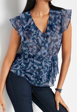 Blouse bleue à motifs sans manches avec un col en V à volants et une ceinture à la taille, associée à un jean bleu foncé et une montre à bracelet bleu.