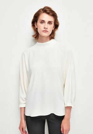 Witte blouse met lange mouwen en hoge hals, zachte textuur en gathered manchetten; comfortabele pasvorm met een vloeiende silhouette. Combi met donkere broek.