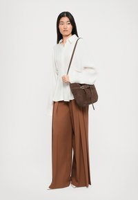 Brown suede crossbody bag with a silver-tone buckle detail. Worn with a white oversized shirt and brown wide-leg trousers.